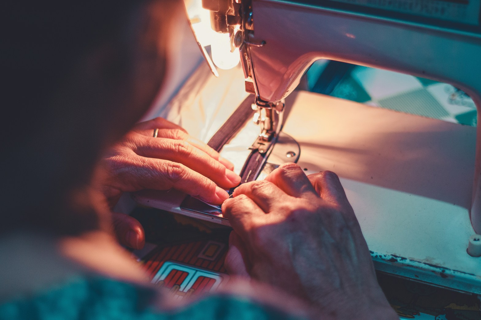 Hands that show signs of age feeding fabric through a sewing machine, taken from the perspective of someone looking over the shoulder of the textile worker