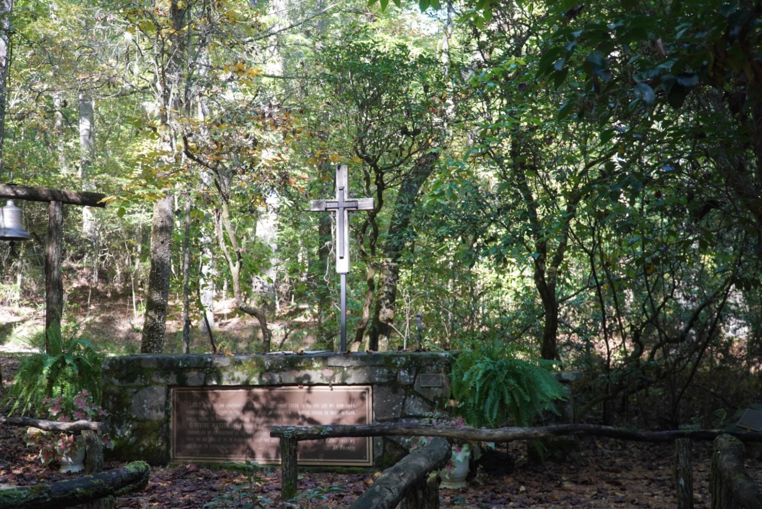 A photo of an outdoor chapel in late summer greens and early fall yellows, with dappled sunlight peaking through